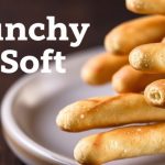 Golden crunchy and soft Breadsticks stacked on a wooden board, sprinkled with herbs.